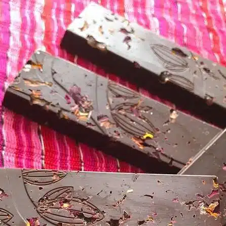 Three dark chocolate bars with flower petal embellishments rest on a vibrant pink and red striped fabric, reminiscent of the colorful charm you'll find exploring Things To Do in Puerto Viejo. The chocolates have decorative patterns engraved on them.