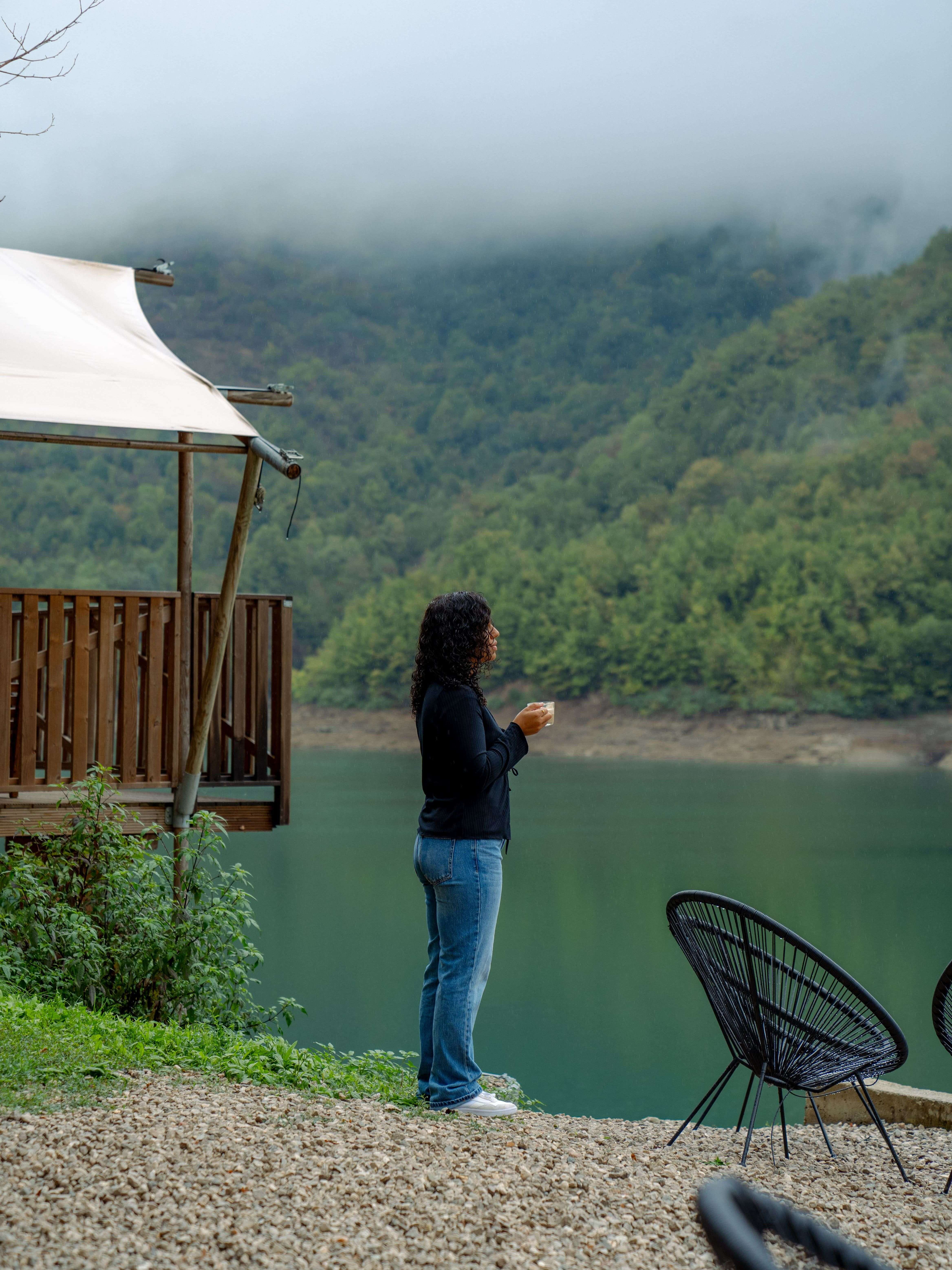 A person stands beside a serene lake enveloped by lush green hills and mist, as if savoring 3 Days in Bosnia and Herzegovina. They hold a cup, dressed casually. Nearby are black chairs and a wooden structure with a canopy, adding to the calm and peaceful atmosphere.