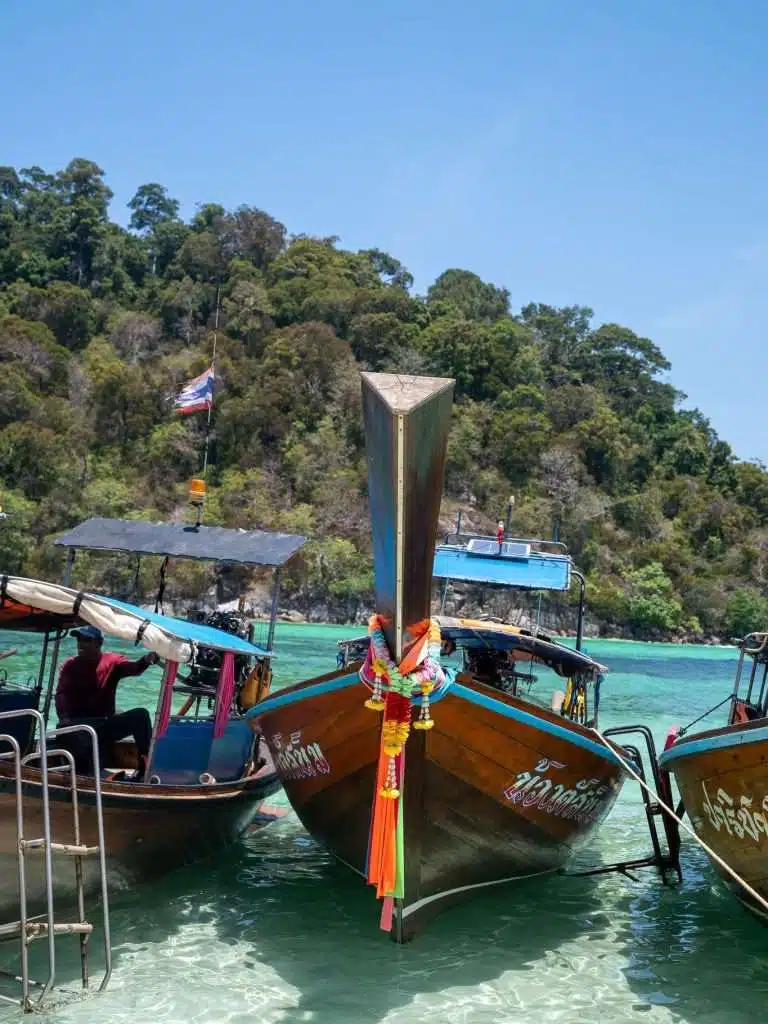 Three traditional wooden long-tail boats with colorful decorations are anchored near a clear turquoise shoreline, one of the exquisite sights among the things to do in Koh Lipe. Lush green trees cover the background under a bright blue sky, embodying tropical paradise.