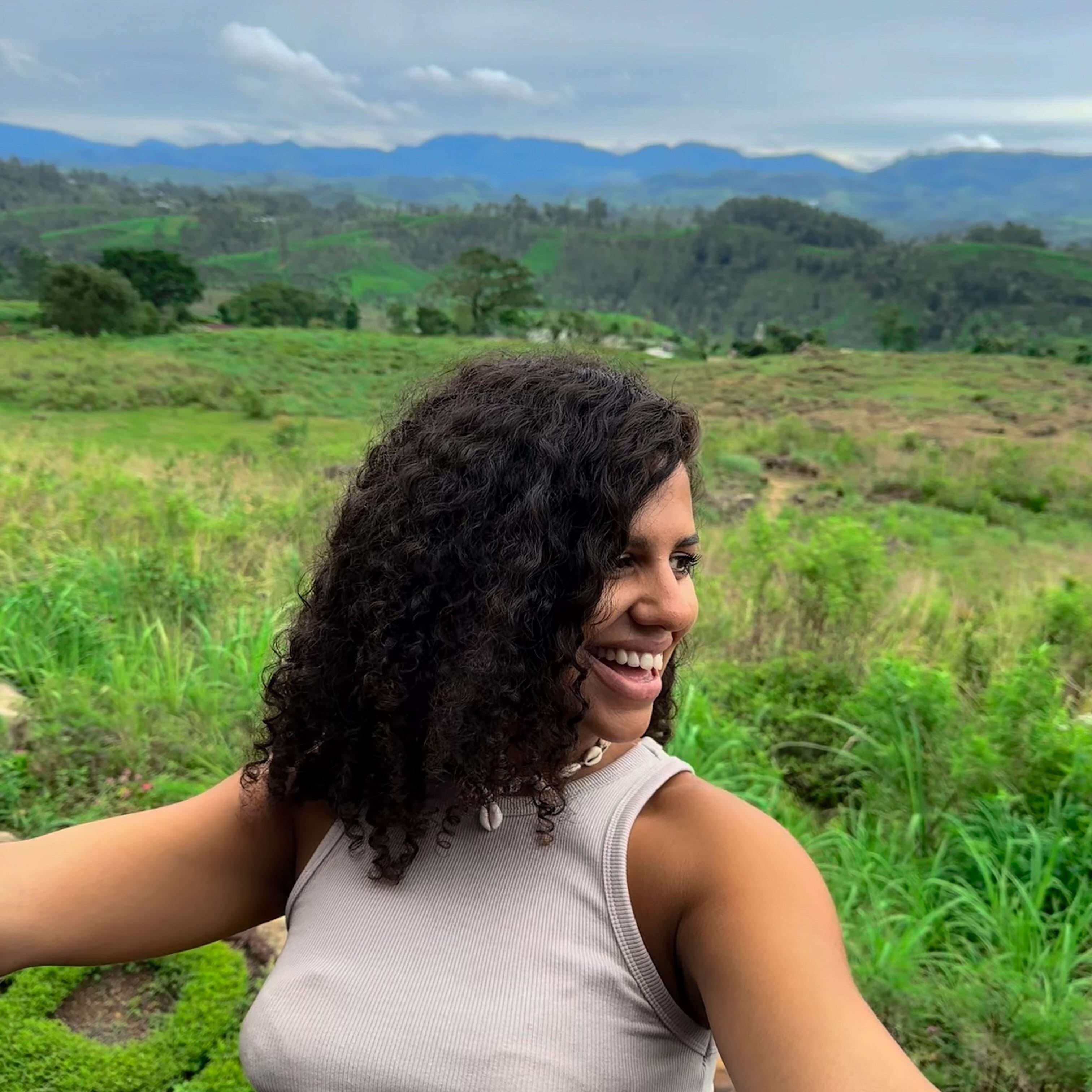 A woman with curly hair smiles while taking a selfie in a lush, green landscape. As she captures the rolling hills and cloudy sky, she dreams of exploring the vibrant restaurants in Ahangama that offer both delightful cuisine and stunning views just like this one.