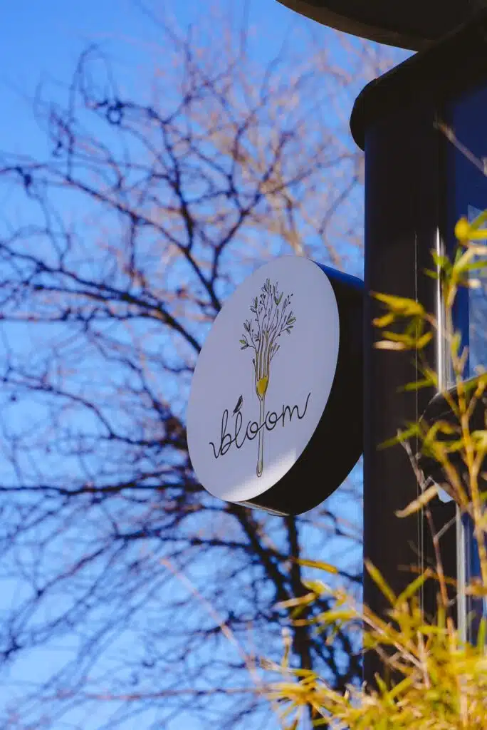 A round sign with an illustration of flowers above the word "bloom" is mounted on a building in Belgrade. Set against a backdrop of a clear blue sky and bare tree branches, it captures the essence of one day in Belgrade, while green plants flourish in the foreground.