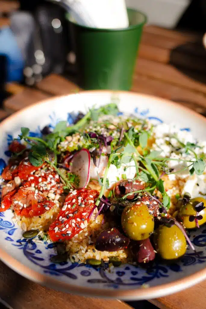 A colorful salad in a white and blue bowl on a wooden table, capturing the essence of one day in Belgrade, features greens, radishes, olives, roasted red peppers, and grains topped with seeds. A blurred green cup sits in the background as bright sunlight enhances the fresh ingredients.
