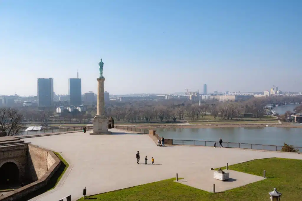 Elevated view of Belgrade Fortress with the iconic Victor Monument overlooking the river, capturing the essence of one day in Belgrade. A few people stroll across the open plaza, while the city skyline and lush trees create a picturesque backdrop under a clear blue sky.