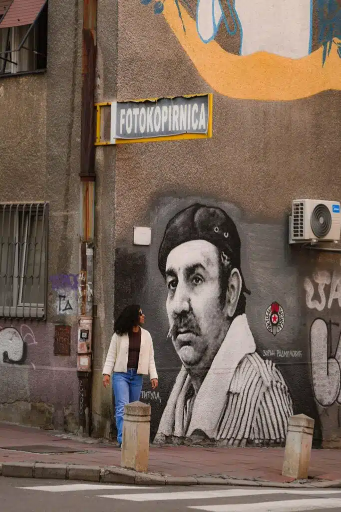 A woman with curly hair and a long coat strolls by a mural on a building wall in Belgrade. The artwork shows a man in a beret and scarf beneath the sign "Fotokopirnica." Air conditioning units are mounted above, capturing just one day in Belgrade's vibrant street scene.