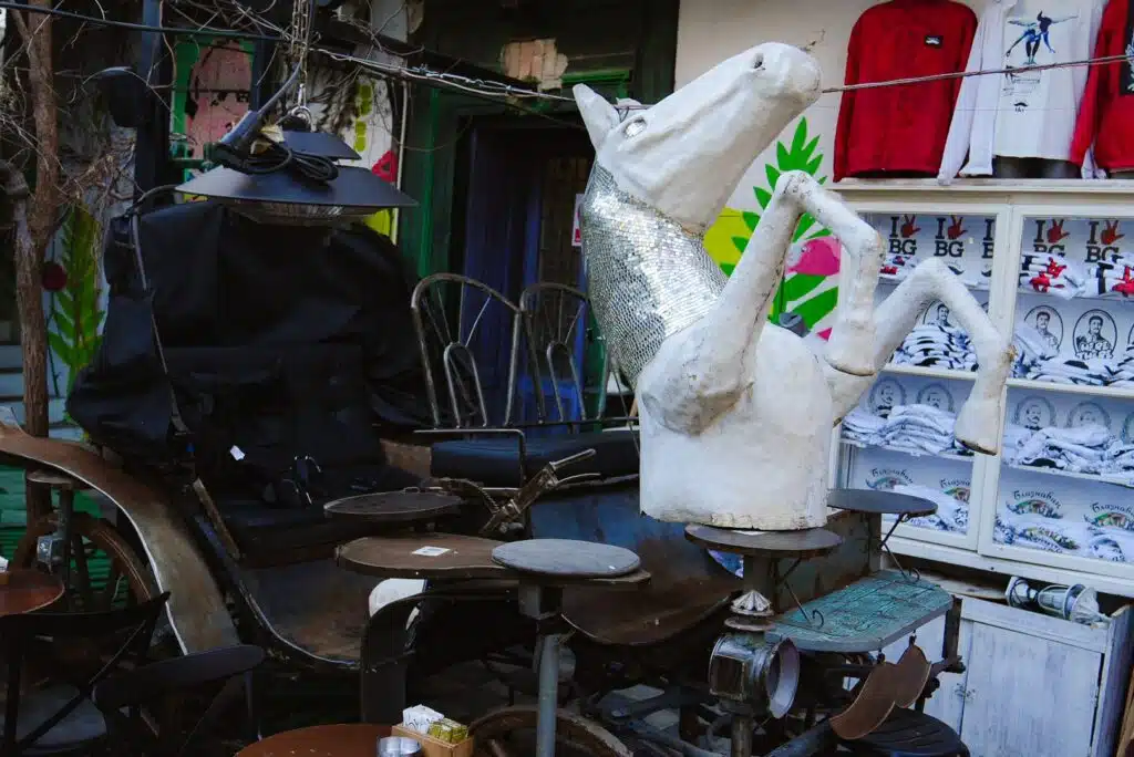 One day in Belgrade, explore an outdoor scene with a vintage carriage, a white horse sculpture draped in a silver scarf, and eclectic decor. T-shirts hang on a wall amidst vibrant murals, creating a quirky and artistic atmosphere.