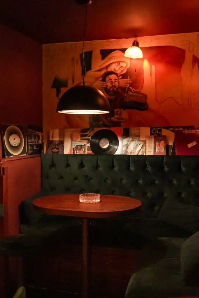 Cozy, dimly-lit corner reminiscent of one day in Belgrade. A round wooden table and tufted seating create a snug atmosphere, while vinyl records and album covers are artfully displayed behind. A large mural and pendant lights enhance the warm ambiance, with an ashtray quietly resting on the table.