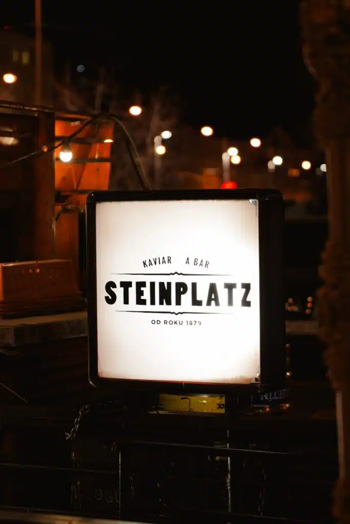 As night falls, an illuminated sign declares "STEINPLATZ," framed by "KAVÍÁR A BAR" above and "OD ROKU 1879" below. The scene captures one day in Bratislava, with dimly lit surroundings and the soft blur of city lights painting the backdrop.