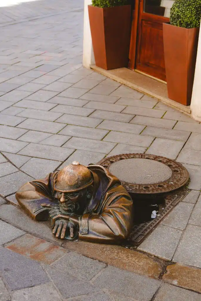 In Bratislava, a bronze statue depicts a man in a hard hat emerging from a manhole on the cobblestone street. With folded arms resting on the ground, he's surrounded by two large planters and a building entrance—an iconic sight for anyone spending one day in this charming city.