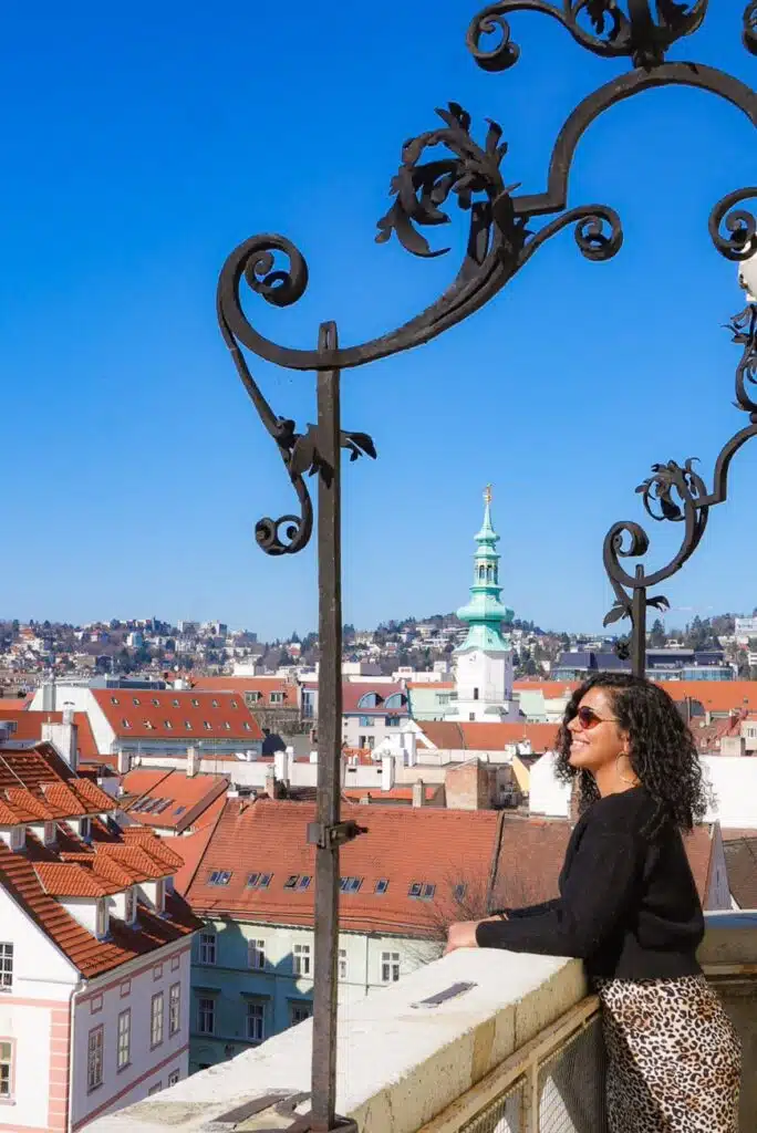 A woman with curly hair and sunglasses leans on a balcony railing, taking in the vibrant rooftops of Bratislava. Beyond, a distant church with a green spire punctuates the clear blue sky, while ornate metalwork frames her view of the cityscape on this perfect day.