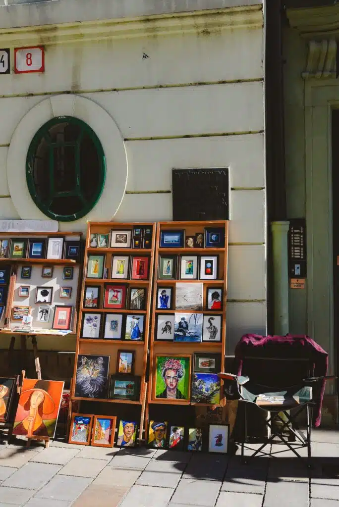 One day in Bratislava, an outdoor art display unfolds with framed paintings and photographs leaning against a building wall. A green circular window presides over the eclectic collection of animals, abstract designs, and portraits. A folding chair stands ready for contemplation.