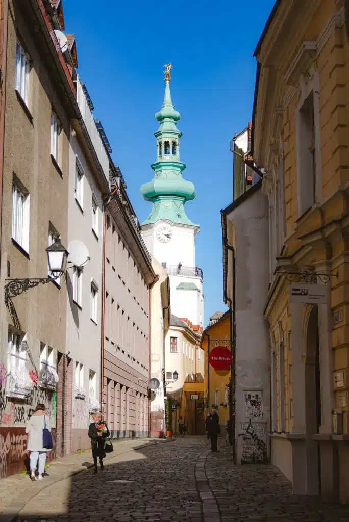 A narrow cobblestone street in Bratislava leads to a tall building with a green spire and clock tower under a blue sky. The street is lined with historic buildings, a few pedestrians, and vintage-style street lamps, capturing the essence of one day in this charming city.