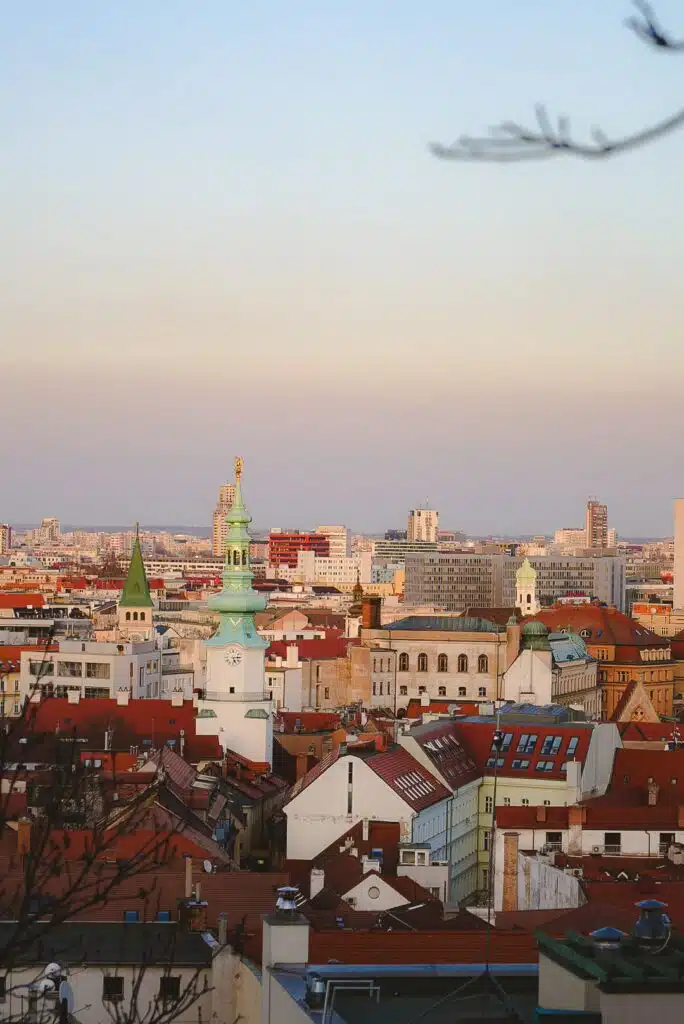 As sunset envelops Bratislava, the cityscape comes alive with a blend of historic and modern buildings crowned by red rooftops. A green-topped tower stands proudly among the dense structures, while the sky gracefully shifts from blue to orange and pink hues for one magical day.