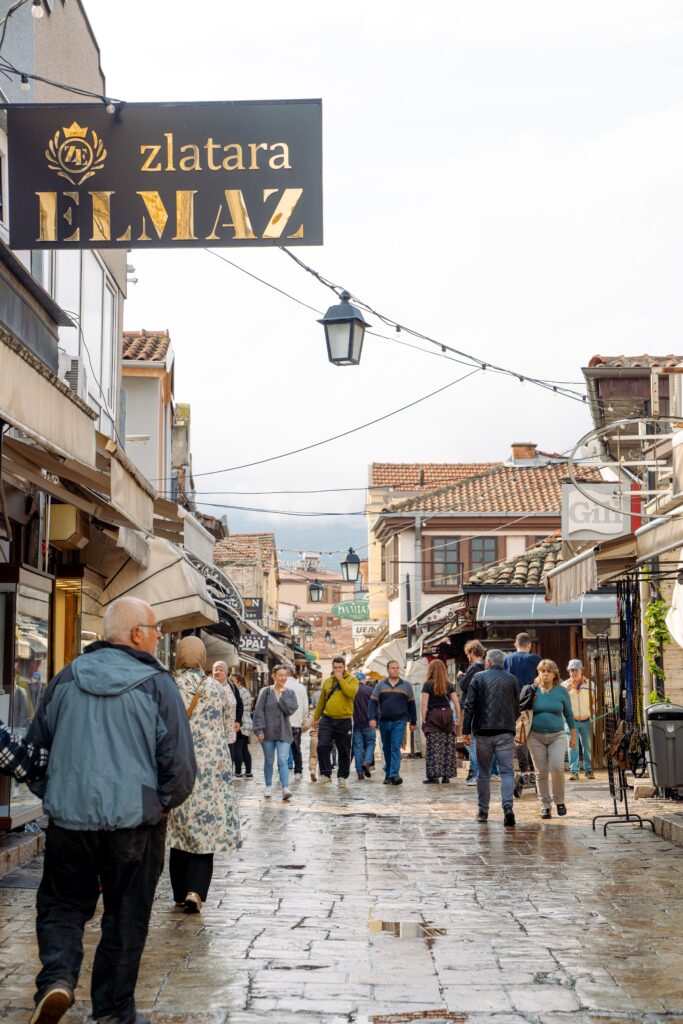On a bustling outdoor market street in Skopje, people stroll along wet cobblestones. Shops line both sides with vibrant signs and enticing goods. A prominent sign reads "Zlatara Elmaz." The sky is overcast, but the atmosphere remains lively for those spending one day in Skopje.