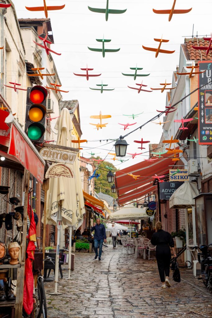 A narrow cobblestone street in Skopje is lined with shops and cafes. Colorful model planes hang above as people stroll along, with a traffic light visible on the left. The setting is lively, a vibrant scene weaving together reds and yellows that captures the essence of one day in this city.