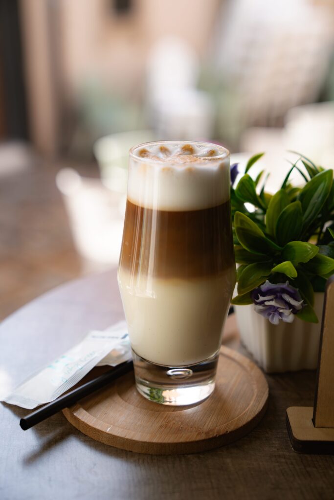 A tall glass of layered iced coffee with milk sits on a wooden coaster, capturing the essence of one day in Skopje. A small plant with green leaves and purple flowers accompanies it. Nearby, a napkin and straw await use, while the background is softly blurred.