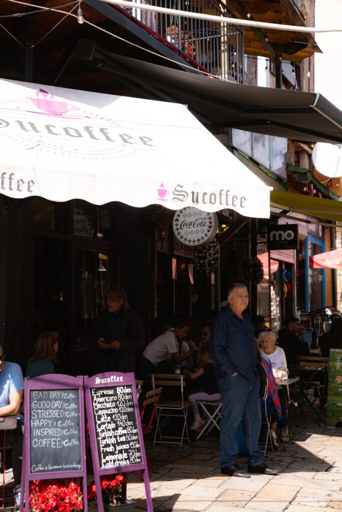 People relax at an outdoor cafe under a white awning with "Sรผcoffee" on it, enjoying the lively scene of one day in Skopje. A man stands nearby while others chat at tables. Two menu boards display drinks and prices, adding to the warm, sunny atmosphere.