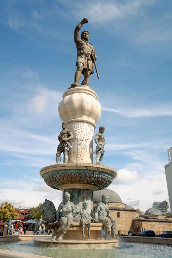 In Skopje, a grand ornamental fountain features a statue of a man with a raised arm atop a carved pedestal. Two smaller statues flank its base, while four sitting figures encircle the water basin. The sky overhead is bright and partly cloudy, adding to this majestic sight.