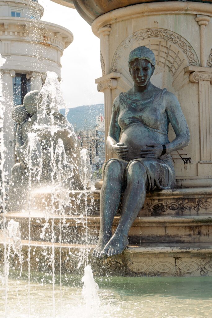 In Skopje, discover a bronze statue of a seated pregnant woman holding her belly, nestled at a fountain. Water splashes in the foreground while intricate carvings embellish the base. Another statue peeks from behind, adding depth to this artistic oasis.