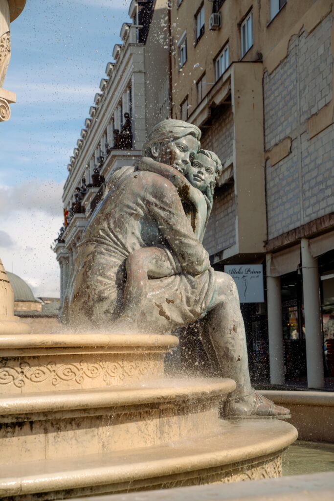 Bronze statue of a man sitting and holding a child on his lap at a fountain in Skopje. Water splashes around them, framed by old buildings under a cloudy sky, capturing a moment from one day in this historic city.