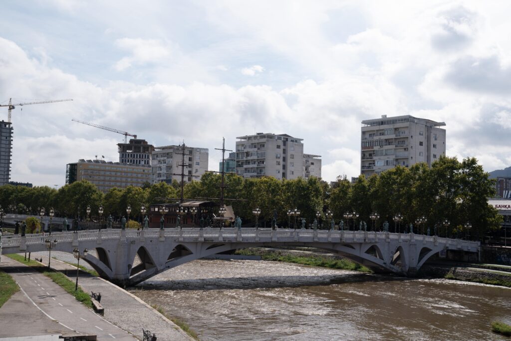 A stone bridge spans a river under cloudy skies, capturing the essence of one day in Skopje. Modern buildings and a line of trees stand proudly in the background. Pedestrians amble on the bridge, while cranes silhouette against the skyline to the left.