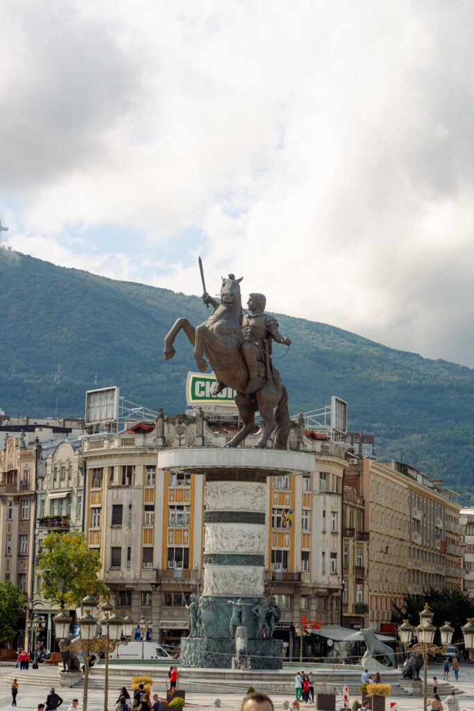 A statue of a warrior on a rearing horse, majestically perched atop a large columned pedestal, stands against the mountainous backdrop. This lively city square, with bustling people and surrounding buildings, offers a perfect glimpse into one day in Skopje beneath the partly cloudy sky.