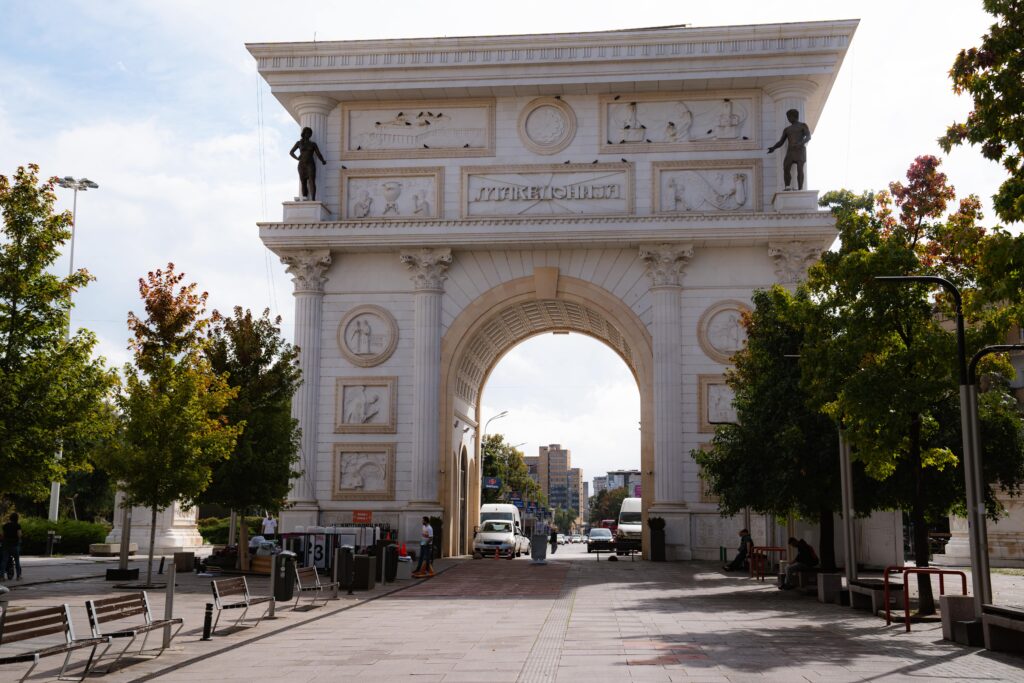 In the heart of Skopje, a large, ornate arch adorned with sculptures and carvings graces the plaza. Surrounded by trees and benches, it's a perfect spot to start your one-day adventure. Through its majestic archway, people and vehicles bustle about on this sunny day.