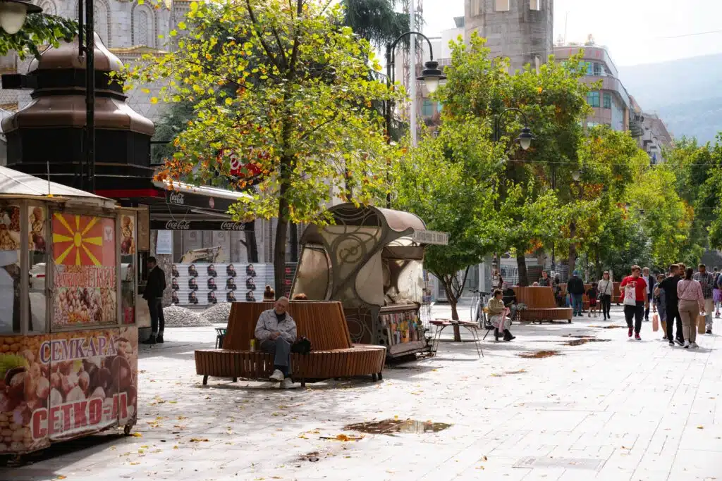 A street scene in Skopje with people strolling and sitting on benches along a tree-lined pedestrian walkway. Food kiosks with signage line the left, while fallen leaves cover the ground. Buildings rise in the background under a cloudy sky, encapsulating one day in this vibrant city.