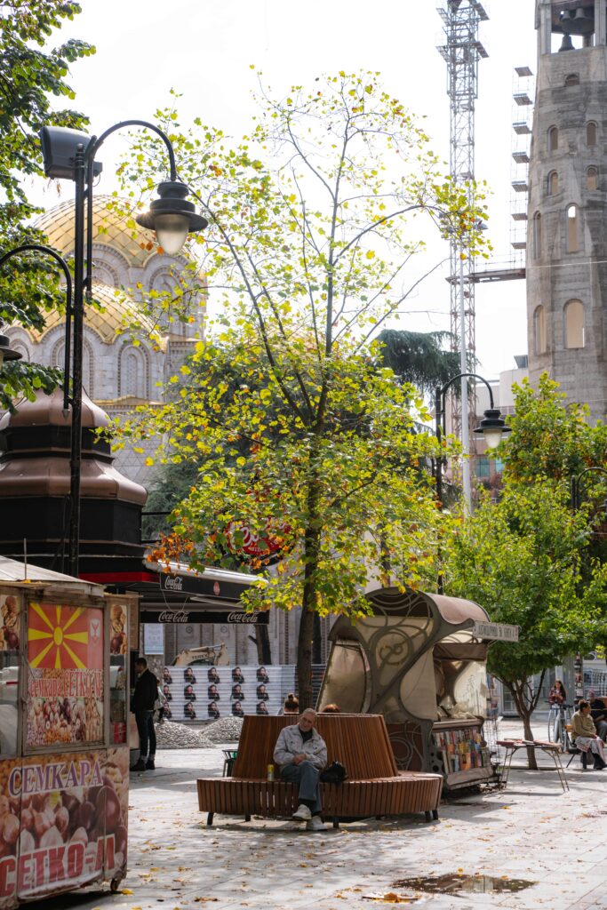 A person sits on a bench in a tree-lined street of Skopje, where a decorative kiosk stands nearby. Behind them, the gold-domed building gleams under autumn's bright leaves scattered on the ground. Street lamps cast gentle light while a construction crane quietly completes this one day in Skopje.