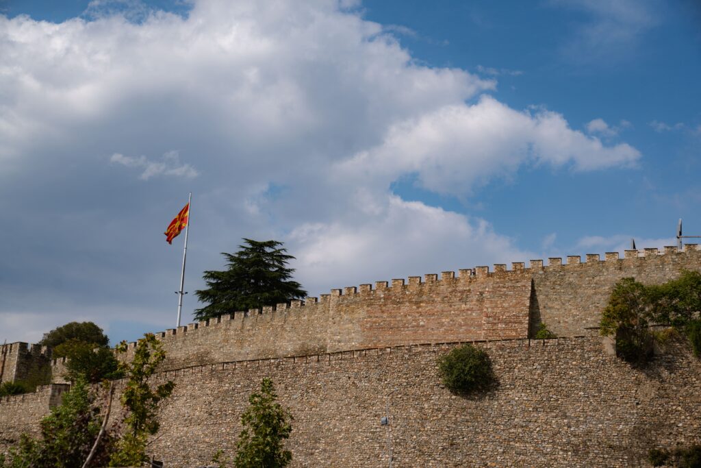 In Skopje for the day, admire stone castle walls with crenellations, crowned by a red and yellow flag waving proudly. Lush trees and greenery frame this majestic view under a blue sky dotted with scattered clouds.