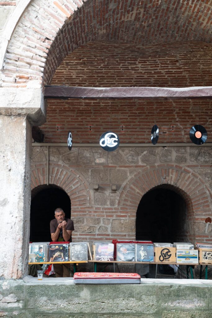 A man leans on a table under an arched brick structure, enveloped by vinyl records hanging above and displayed below. This unique outdoor scene, reminiscent of one day in Skopje, is framed by stone walls and two arched openings.