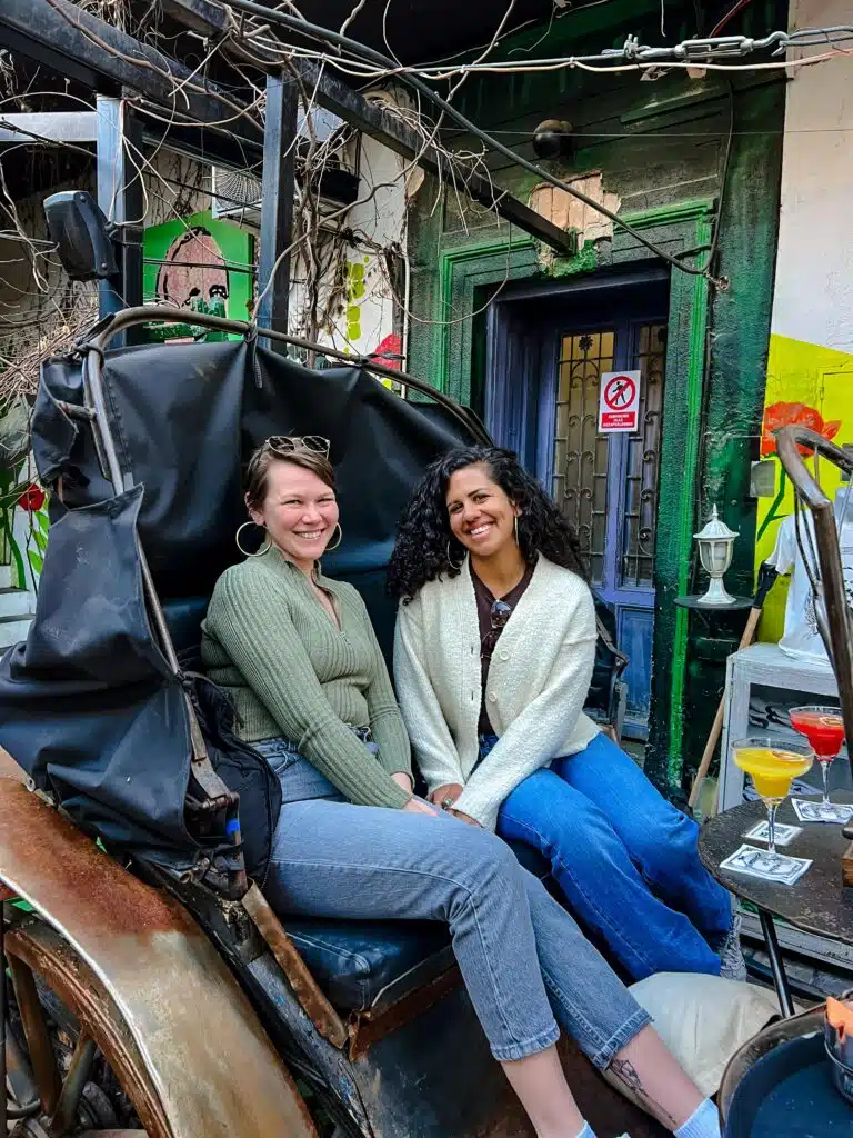 Two people are sitting on a vintage carriage seat outdoors, enjoying one day in Belgrade. Both are smiling; one wears a green sweater and the other a white cardigan. Beside them is a small table with colorful drinks, set against an eclectic backdrop of a painted wall.