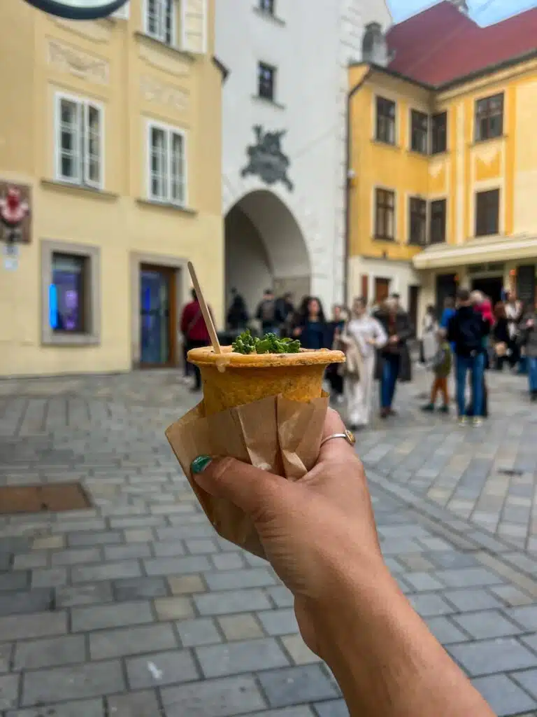 A hand holds a cone of soup garnished with herbs, set against a historic European backdrop in Bratislava. Through the stone archway, people wander along the cobblestone streets, capturing the essence of one day in this charming city.