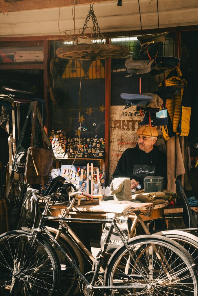 A man sits at a cluttered workbench surrounded by various items and bicycles. He's wearing a cap and glasses, working with tools in a shop filled with hats, buttons, and trinkets. Sunlight streams in through the window, casting a warm glow reminiscent of one day in Skopje.