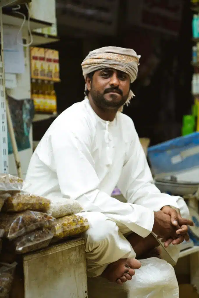 A man wearing a traditional white robe and headscarf sits on a counter in a store, embodying the essence of an enriching 1-week Oman itinerary. Barefoot and surrounded by bags of spices, his clasped hands and calm expression reflect the tranquility found in Oman's vibrant markets.