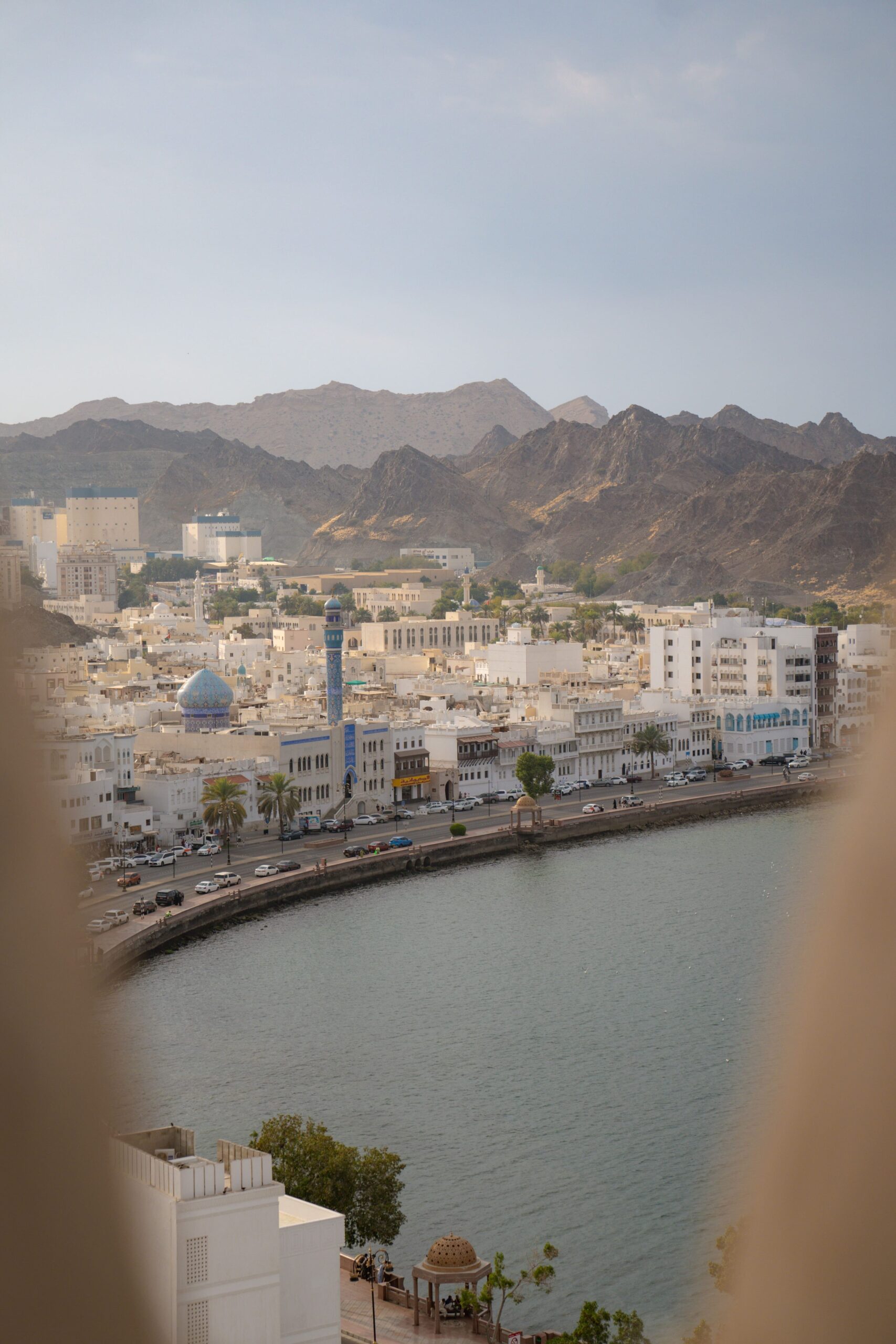 Scenic view of a city with white buildings along a waterfront, surrounded by rugged mountains—a must-see in your 1-week Oman itinerary. A blue minaret stands out among the structures, and an arch frames the serene scene under a clear sky.