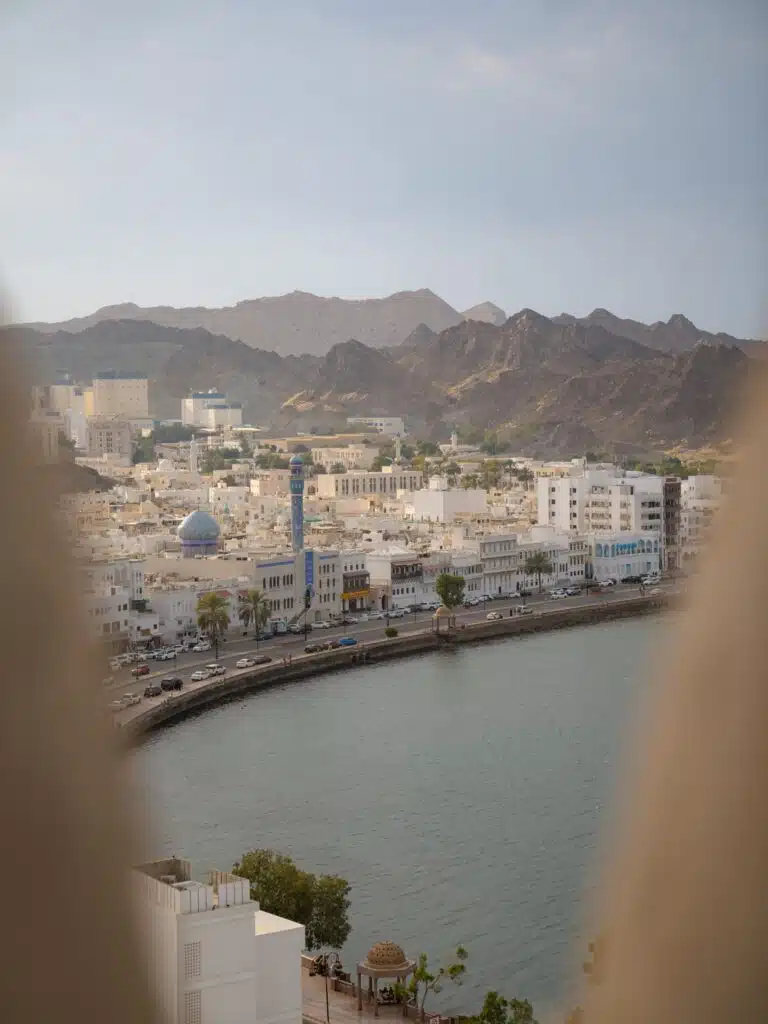 Scenic view of a city with white buildings along a waterfront, surrounded by rugged mountains—a must-see in your 1-week Oman itinerary. A blue minaret stands out among the structures, and an arch frames the serene scene under a clear sky.
