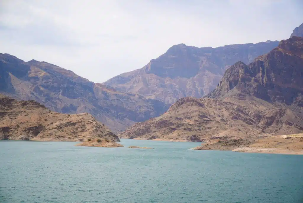 A serene lake with turquoise waters is surrounded by rugged mountains under a partially cloudy sky. The landscape, reminiscent of a 1-week Oman itinerary, features rocky hills and varied terrain, creating a peaceful and remote natural scene.