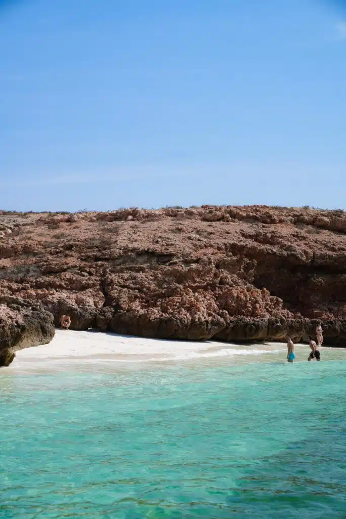 Clear turquoise water meets a sandy beach bordered by rocky cliffs under a bright blue sky, reminiscent of a perfect stop on a 1-week Oman itinerary. Two people stand in the shallow water, enjoying the serene coastal scene.