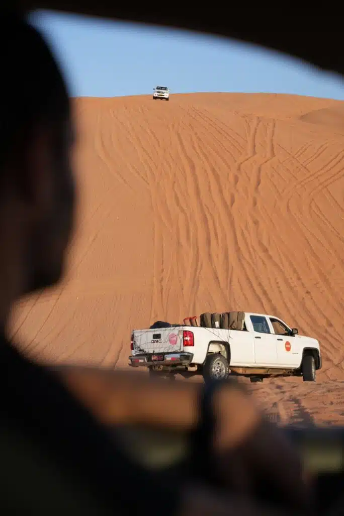 A pickup truck with a spare tire in the back and Husaak Adventures written across it is parked on a sandy dune, reminiscent of scenes one might encounter during a 1-week Oman itinerary. Another vehicle is visible atop the dune. In the foreground, a blurry figure sits, observing under the expansive clear blue sky.