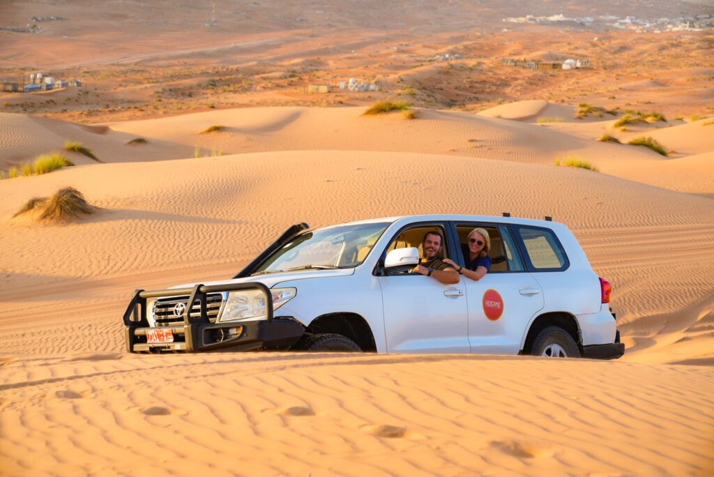 A white SUV glides through the vast desert of a 1-week Oman itinerary, weaving between majestic sand dunes. Inside, a smiling couple takes in the breathtaking views on this bright, sunny day. Sparse vegetation and distant structures add intrigue to their unforgettable adventure.