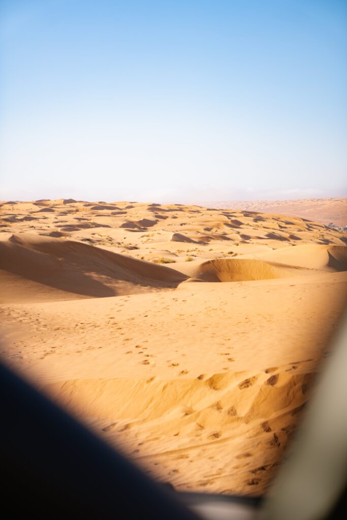 Expansive desert landscape with golden sand dunes under a clear blue sky, perfect for your 1-week Oman itinerary. The sun casts shadows on the dunes, creating patterns on the sand. Out-of-focus elements frame the foreground, enhancing the sense of depth.