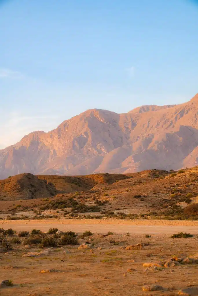 Experience the enchanting beauty of Oman with a 1-week itinerary, where sunlit mountains rise majestically against a rocky desert landscape under a clear blue sky.