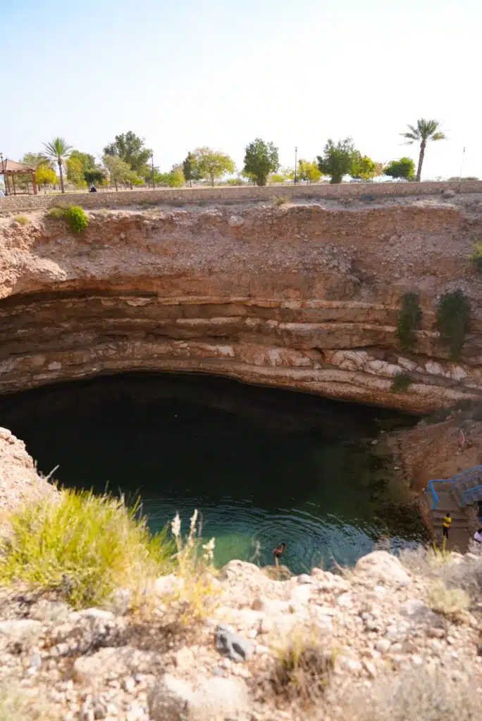 A natural sinkhole filled with clear blue water is a must-see on any 1-week Oman itinerary. Surrounded by rocky walls, the site features sparse vegetation and a few swimmers enjoying the refreshing waters. In the background, palm trees and a fence line stand out against a bright sky.