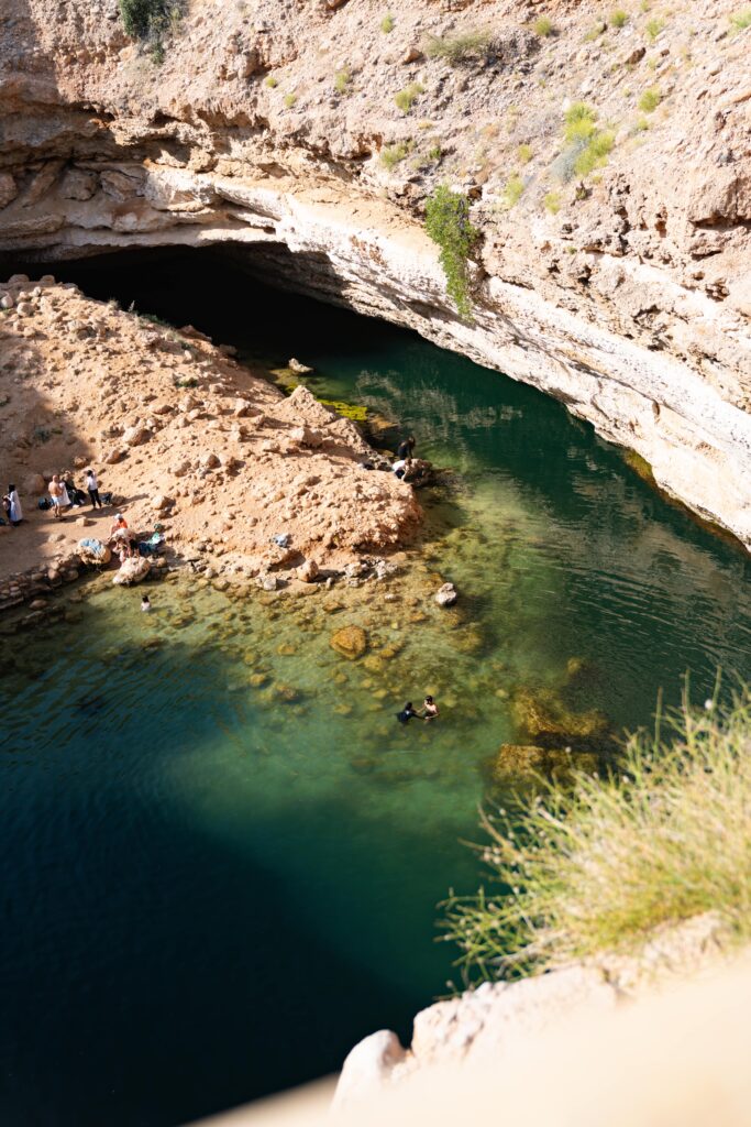 Nestled as a must-visit on a 1-week Oman itinerary, this natural limestone sinkhole is filled with clear green water and encircled by rocky cliffs. A few people enjoy swimming and relaxing at the water's edge, where bright sunlight casts playful shadows across the rugged landscape.
