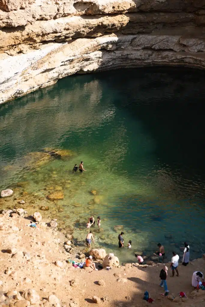 Aerial view of people swimming and relaxing in a natural water pool surrounded by rocky cliffs, reminiscent of stops on a 1-week Oman itinerary. The clear water transitions from turquoise to deeper shades of green as visitors gather on the sandy shore and swim in the pool.