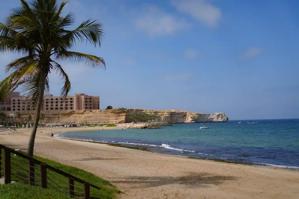 Amidst a serene beach scene with a sandy shore, a solitary palm tree stands tall by the clear blue ocean. A few small boats dot the water, while a large pink building overlooks from a rocky cliff. This picturesque spot is an ideal beginning for your 1-week Oman itinerary under a partly cloudy sky.