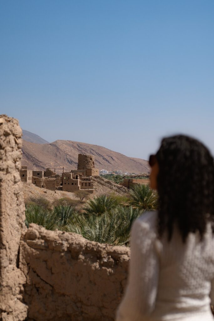 A person with long curly hair and sunglasses gazes over an ancient Omani desert village, nestled among palm trees and mud-brick buildings, with a mountain range in the background stretching into the clear blue sky—a perfect start to a 1-week Oman itinerary.