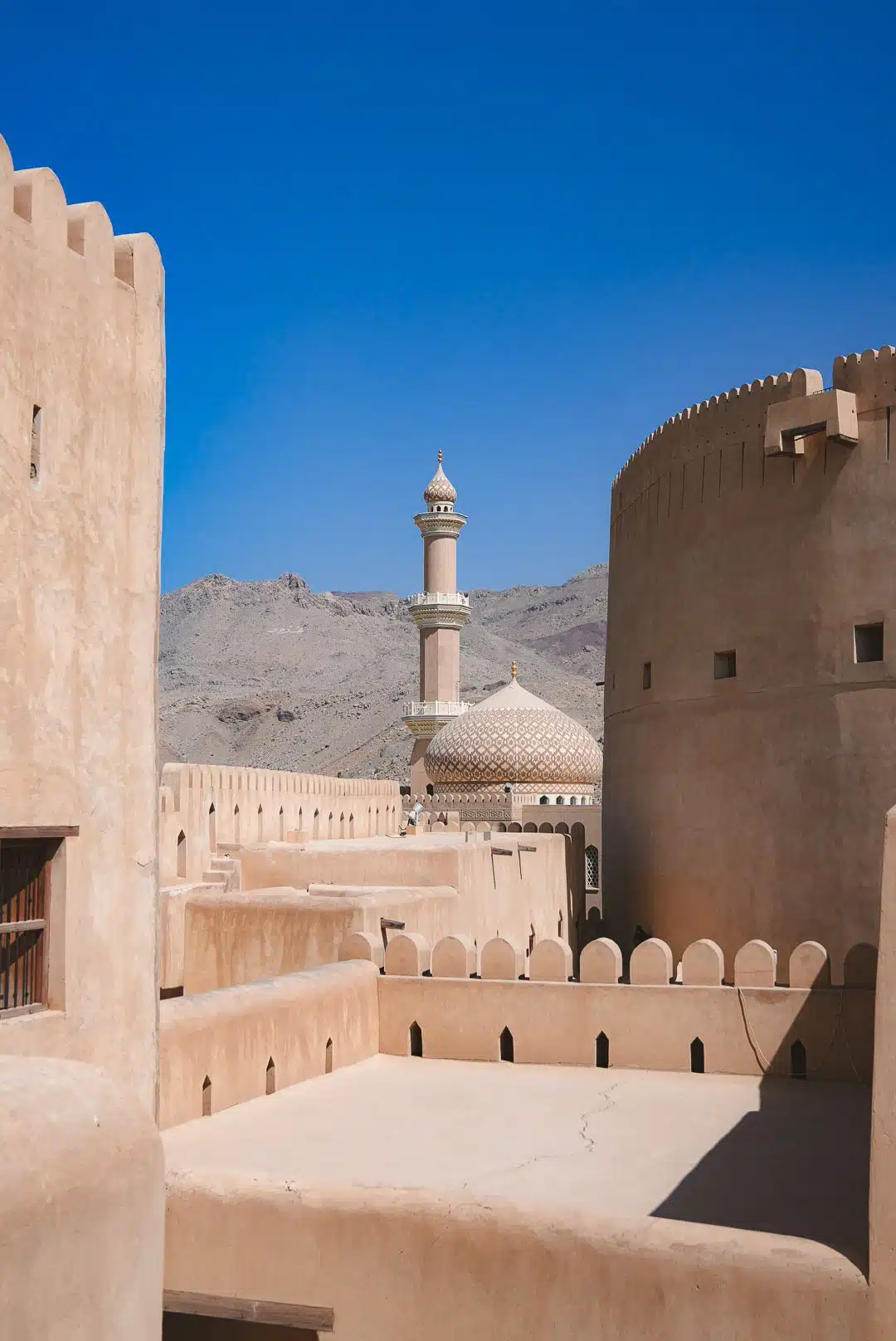 Historic desert fort with sand-colored stone walls, topped with crenellations. A distant mosque with a dome and minaret is visible against a backdrop of arid mountains and a clear blue sky—must-see destinations on your Oman itinerary.