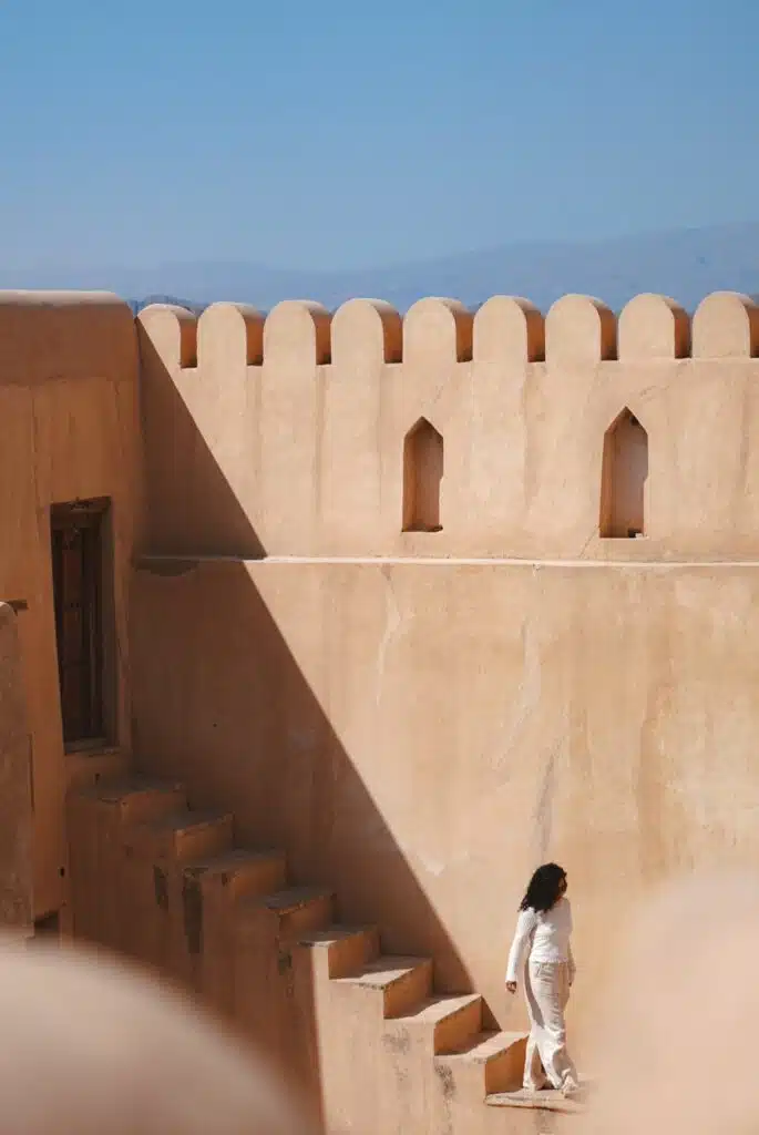 A person in white clothing ascends a staircase in an ancient, sandy-colored fort—possibly a highlight on a 1-week Oman itinerary. The scene is bathed in sunlight under a clear blue sky, with distant mountains nearby.