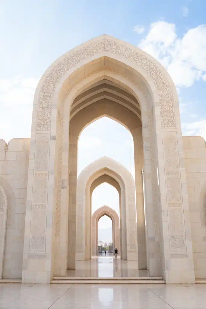 An intricate series of white stone archways with ornate carvings leads into the distance under a blue sky with light clouds, reminiscent of a journey through time on a 1-week Oman itinerary. The polished floor reflects the arches, creating a perfectly symmetrical view.